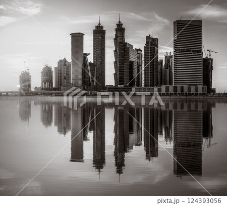 Monochrome city skyline with tall skyscrapers reflecting on calm water under a cloudy sky, Dubai Marina bay UAE 124393056