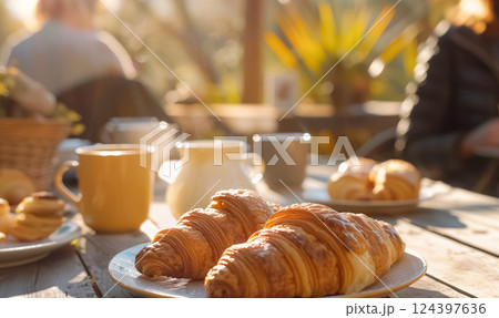 Fresh croissants on breakfast table in warm morning sunlight, people gathered in the background, creating cozy, inviting atmosphere for brunch or coffee or tea 124397636