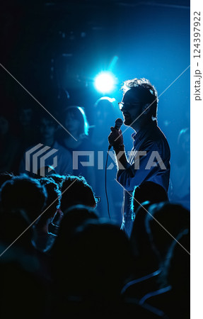 silhouette of man person holding microphone on stage, backlit by blue light. The crowd watches in anticipation, creating a dramatic and intense atmosphere 124397922