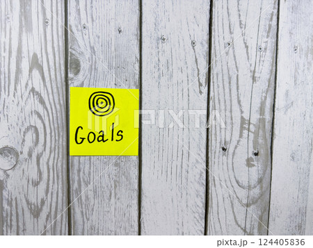 Sticky note showcasing the word Goals accompanied by target symbol, set against textured wooden background, symbolizing the importance of setting and achieving personal aspirations Sticky note showcasing the word Goals accompanied by target symbol, set against textured wooden background, symbolizing the importance of setting and achieving personal aspirations 124405836