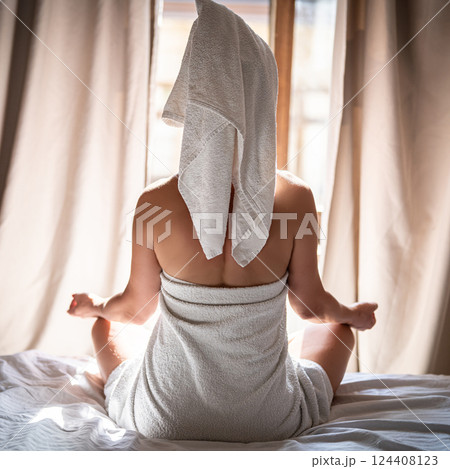 Back view of young woman in towel sitting on bed and meditating Back view of young woman in towel sitting on bed and meditating 124408123