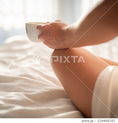 Cropped image of woman holding cup of coffee while sitting on bed at home 124408141