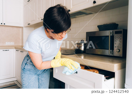 Mature female cleaning kitchen drawer with gloves and cloth 124412540