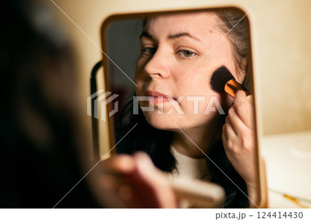 Woman doing make up looking in mirror at home. Putting on make-up with brush. Applying blush to face. Reflection in mirror. Woman doing make up looking in mirror at home. Putting on make-up with brush. Applying blush to face. Reflection in mirror. 124414430