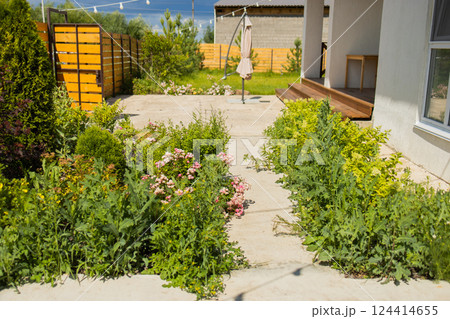 Concrete walkway between flowerbeds leads to wooden gate. Structured garden planning and decorative landscape in residential space. Concrete walkway between flowerbeds leads to wooden gate. Structured garden planning and decorative landscape in residential space. 124414655
