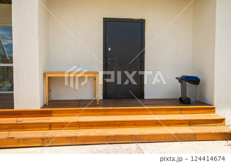 Black entrance door with trash bin and wooden table. Neat home access and minimalistic outdoor arrangement for everyday use Black entrance door with trash bin and wooden table. Neat home access and minimalistic outdoor arrangement for everyday use 124414674