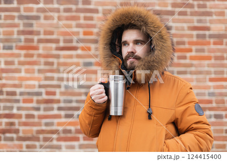 Man with thermos stands by brick wall in winter street. Urban lifestyle, seasonal routine, and outdoor relaxation in cold weather. Man with thermos stands by brick wall in winter street. Urban lifestyle, seasonal routine, and outdoor relaxation in cold weather. 124415400