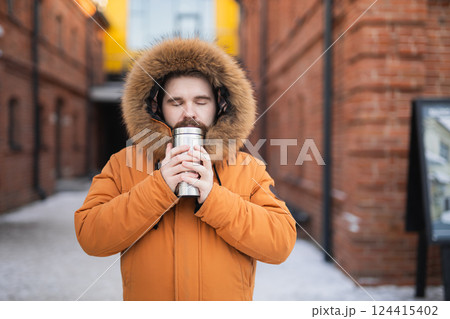 Man in winter parka holds thermos with closed eyes on snowy urban street. Warmth, solitude, and comfort in cold weather. Man in winter parka holds thermos with closed eyes on snowy urban street. Warmth, solitude, and comfort in cold weather. 124415402