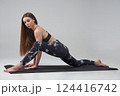 Barefoot Caucasian girl doing lizard position of yoga in studio. Side view of pretty female in activewear looking at camera, while practicing yoga on mat, isolated on grey background. Yoga concept.  124416742
