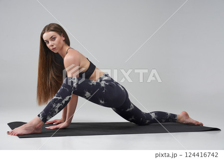 Barefoot Caucasian girl doing lizard position of yoga in studio. Side view of pretty female in activewear looking at camera, while practicing yoga on mat, isolated on grey background. Yoga concept.  124416742