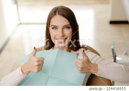 Smiling beautiful girl is sitting in dentistry chair with positive emotion and gesturing fingers up. Female patient visit dentist for regular teeth mouth cavity check up at medical clinic 124416853