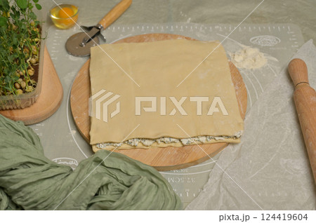 Square sheet of dough on wooden board. Rolling pin, cutter, egg, green towel, microgreens sprouts on kitchen table. Home cooking baking puffs Square sheet of dough on wooden board. Rolling pin, cutter, egg, green towel, microgreens sprouts on kitchen table. Home cooking baking puffs 124419604