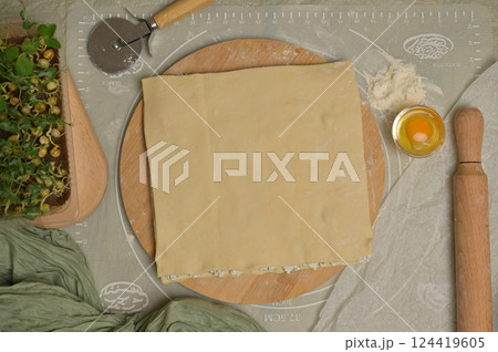 Top view square sheet of dough on wooden board. Rolling pin, cutter, egg, green towel, microgreens sprouts on kitchen table. Home cooking baking puffs 124419605