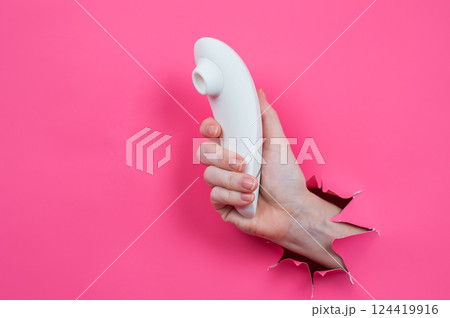 Female hand sticks out white vacuum clitoral vibrator through pink paper background. Female hand sticks out white vacuum clitoral vibrator through pink paper background. 124419916