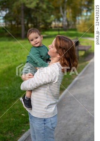 Caucasian woman holds her one-year-old son in her arms while walking in the park in autumn. Caucasian woman holds her one-year-old son in her arms while walking in the park in autumn. 124420225