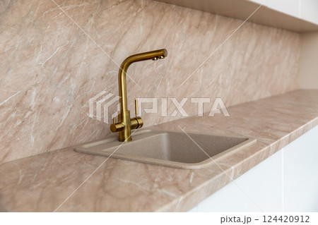A golden faucet in a modern kitchen with a marble beige facade. 124420912