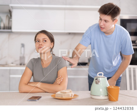 Young girl sitting at the table while her boyfriend is quarreling with her standing behind Young girl sitting at the table while her boyfriend is quarreling with her standing behind 124432838