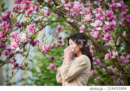 Woman allergic suffering from seasonal allergy at spring in blossoming garden at springtime. Young woman sneezing and blowing nose with nasal handkerchief in front of blooming tree. Allergy concept. Woman allergic suffering from seasonal allergy at spring in blossoming garden at springtime. Young woman sneezing and blowing nose with nasal handkerchief in front of blooming tree. Allergy concept. 124433984