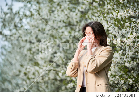 Woman allergic suffering from seasonal allergy at spring in blossoming garden at springtime. Young woman sneezing and blowing nose with nasal handkerchief in front of blooming tree. Allergy concept. 124433991