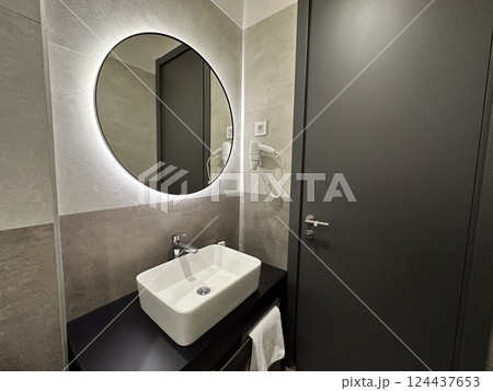Modern bathroom with round mirror and stylish sink in contemporary hotel. Cleanliness, comfort, hygiene, and modern minimalist interior design. Modern bathroom with round mirror and stylish sink in contemporary hotel. Cleanliness, comfort, hygiene, and modern minimalist interior design. 124437653