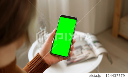 Young woman sitting at home holding smartphone green mock-up screen in hand. Female person use chroma key mobile phone. Vertical mode. Touching, swipe display, tapping, surfing internet social media Young woman sitting at home holding smartphone green mock-up screen in hand. Female person use chroma key mobile phone. Vertical mode. Touching, swipe display, tapping, surfing internet social media 124437779