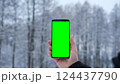 Young man's hand holding smartphone with green screen against snowy forest background. Vertical mockup of mobile phone. Man using phone with blank green display background. Smartphone with chroma key 124437790