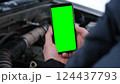 Men's hands are holding a smartphone with a bright green screen against the background of a car engine. Automotive technology. Mobile apps for drivers. Road safety. Using gadgets. Chromakey 124437793