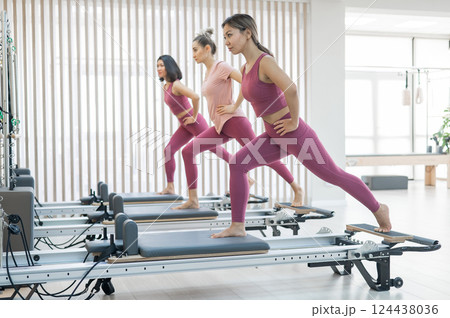 Three Asian women in pink sportswear doing pilates exercises with a reformer bed.  124438036
