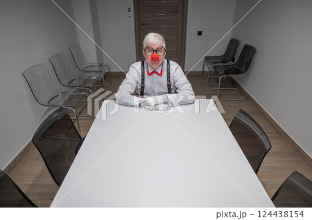 An elderly man in a clown costume sits at a desk in an office.  124438154