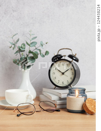 Alarm clock, books, coffee cup, eyeglasses and candle creating cozy atmosphere on wooden desk Alarm clock, books, coffee cup, eyeglasses and candle creating cozy atmosphere on wooden desk 124438214
