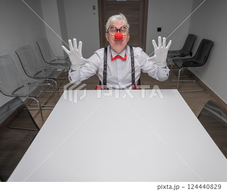 An elderly man in a clown costume sits at a desk in an office.  124440829