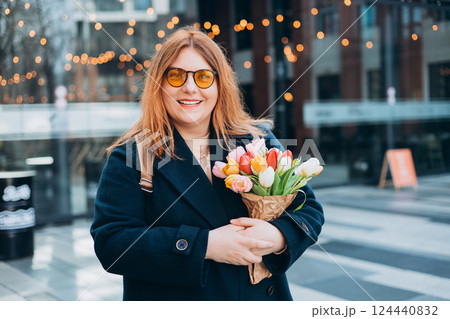 Beautiful young woman with spring tulips flowers bouquet walking at city street. Happy girl posing outdoors. Spring portrait of pretty female on urban background. High quality photo. 124440832