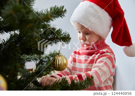 girl in a Santa hat hangs a Christmas ball on a Christmas tree 124440856