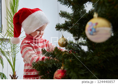 happy little girl in a Santa Claus hat is decorating the Christmas tree with toys and trinkets. Cute baby is decorating home to celebrate Christmas. happy little girl in a Santa Claus hat is decorating the Christmas tree with toys and trinkets. Cute baby is decorating home to celebrate Christmas. 124440857
