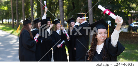 Group of happy students in graduation gowns outdoors. A young girl with a diploma in her hands in the foreground. Group of happy students in graduation gowns outdoors. A young girl with a diploma in her hands in the foreground. 124440938