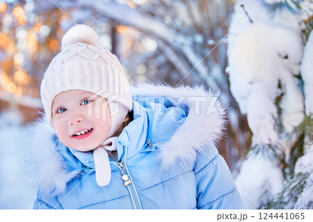 portrait of a smiling child looking into the frame into the camera. Winter landscape with a pine tree in the snow. The baby is dressed in a winter jacket with fur and a warm knitted hat 124441065