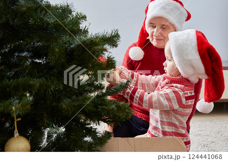 little girl hangs decorations on the Christmas tree. Grandmother admires her granddaughter. The concept of family and New Year's Christmas holidays. little girl hangs decorations on the Christmas tree. Grandmother admires her granddaughter. The concept of family and New Year's Christmas holidays. 124441068