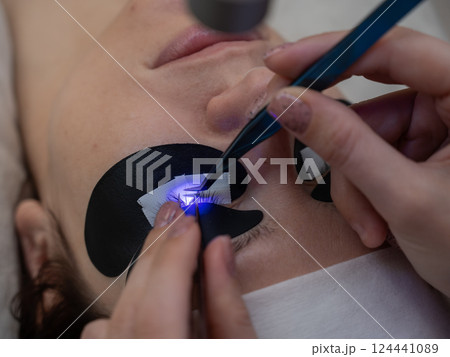 A woman undergoing eyelash extension procedure using an ultraviolet lamp. A woman undergoing eyelash extension procedure using an ultraviolet lamp. 124441089