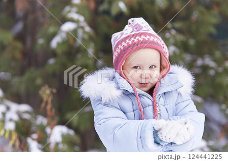 A smiling girl in a pink hat and a blue jacket with fur. In winter, he holds snow in his hands. Looks at the camera. Place for text, banner, new year. 124441225