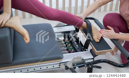 Personal Pilates training. Instructor adjusts woman on reformer.  124441480