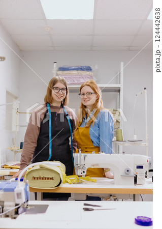 Two female seamstresses in workshop at factory are standing at a table with sewing machines. Two female seamstresses in workshop at factory are standing at a table with sewing machines. 124442638