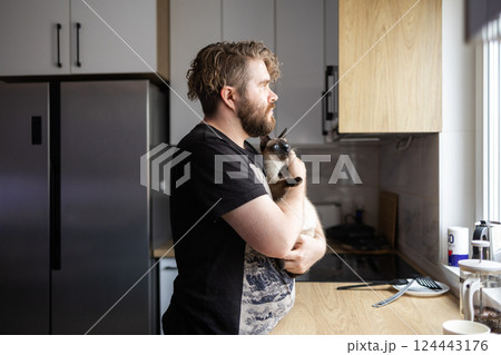 Man holding cat in quiet kitchen moment near window light. Comfort, pet emotional connection, and the peaceful rhythm of everyday domestic life Man holding cat in quiet kitchen moment near window light. Comfort, pet emotional connection, and the peaceful rhythm of everyday domestic life 124443176