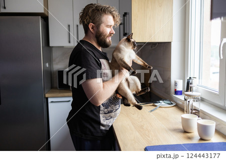 Man holding cat in quiet kitchen moment near window light. Comfort, pet emotional connection, and the peaceful rhythm of everyday domestic life Man holding cat in quiet kitchen moment near window light. Comfort, pet emotional connection, and the peaceful rhythm of everyday domestic life 124443177