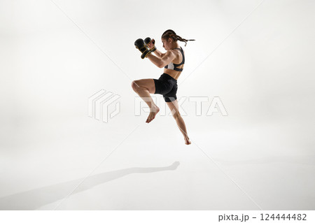 Focused athlete in black boxing gear performing high jump for attack move, training against white background. Focused athlete in black boxing gear performing high jump for attack move, training against white background. 124444482