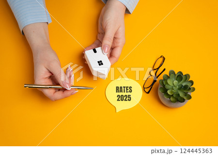 Female hands is a miniature house near notes with the words Rates 2025. Loan and credit interest. Apartments, housing, house. Real estate market analysis concept. Business and Finance 124445363