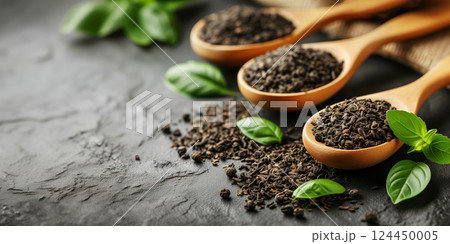 Dry black tea in wooden spoons and green leaves on black background with copy space. 124450005