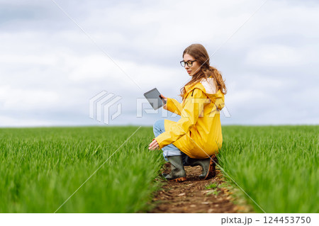 Young farmer woman in a green field, intently using a tablet. She wears a yellow jacket and glasses Young farmer woman in a green field, intently using a tablet. She wears a yellow jacket and glasses 124453750