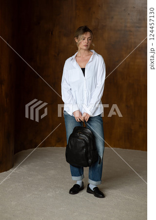 A girl in a white shirt blue jeans and loafers is posing with a backpack. 124457130