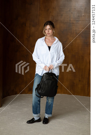 A girl in a white shirt blue jeans and loafers is posing with a backpack. 124457131