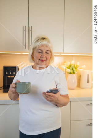 Senior woman holding a phone and a coffee cup in the kitchen, smiling 124458456